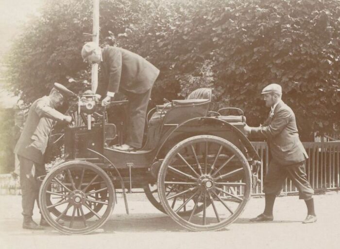 Three men inspecting and pushing a wild first car from 100 years ago with large wooden wheels outdoors.