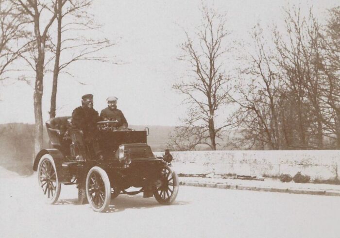 Two men dressed in early 1900s attire driving a wild first car on a rural road with leafless trees around.