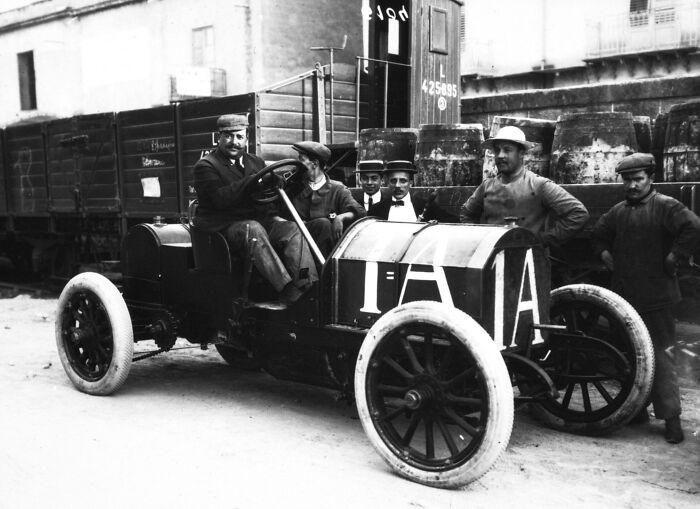 Vintage photo of a wild first car from 100 years ago with men posing around it in a historic urban setting.