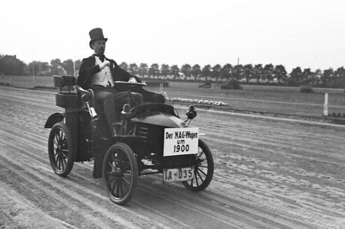 Early 1900s first cars showing wild vintage automobile design with driver in formal attire on dirt road.