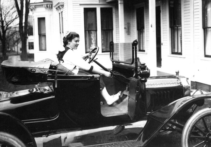 Woman sitting in a wild first car from 100 years ago, showcasing early automotive design in front of a vintage house.