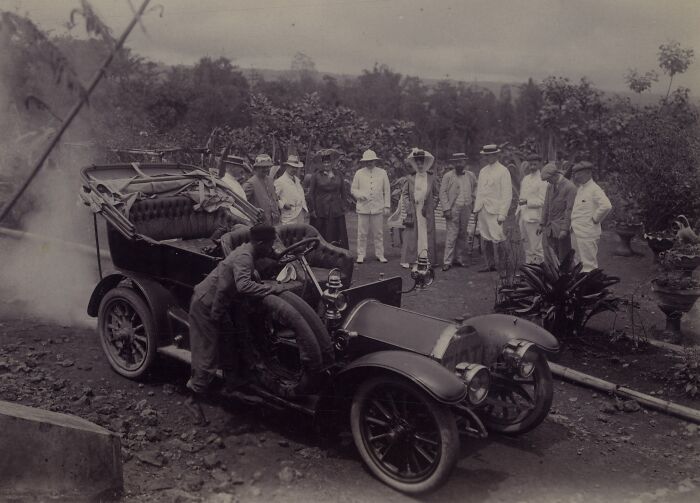Vintage black and white photo showing a wild first car from 100 years ago with people gathered around outdoors.