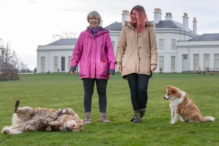 Two women outdoors with dogs, one dog lying playfully on grass, capturing funny times pets crashed photos.