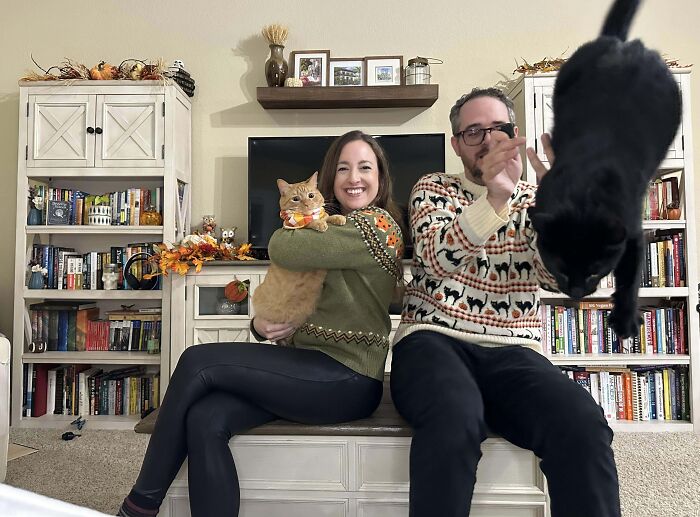 A woman holding a cat smiling while another cat jumps in front, a funny times pets crashed photos moment in a living room.