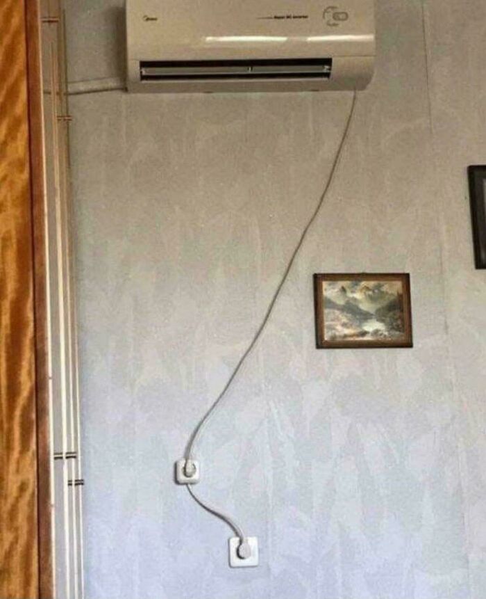 Wall-mounted air conditioner with a long, awkwardly routed power cord plugged into a low wall outlet in a quirky engineering setup.