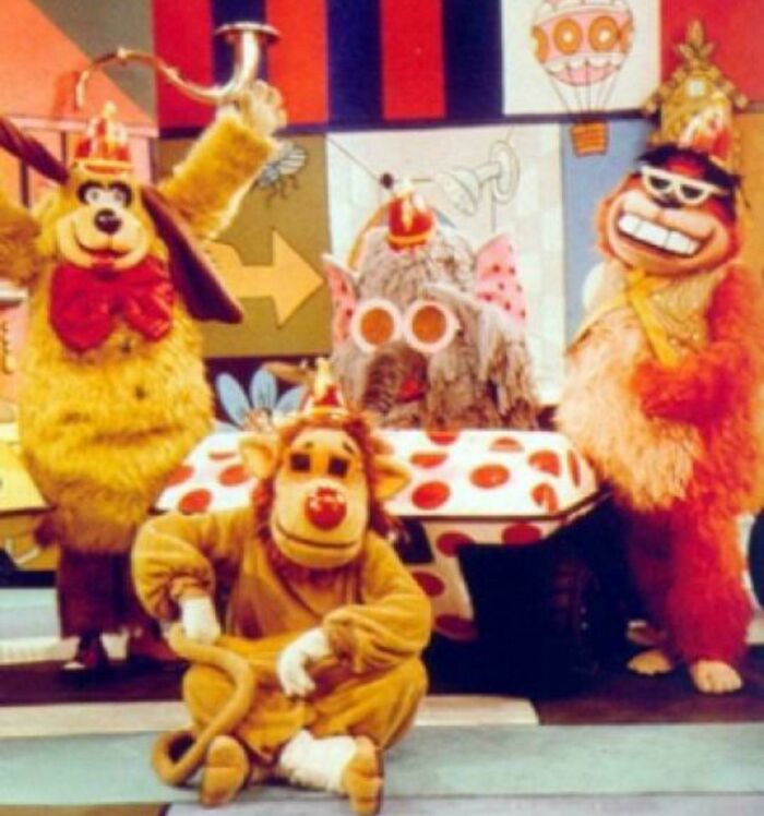 Costumed characters from a vintage children’s TV show pose with bright props, evoking nostalgia to make you feel old.