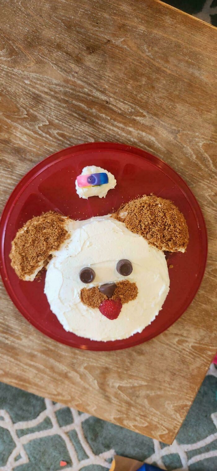 Creative baked goods shaped like a dog face with frosting, cookie crumbs, and candy on a red plate.