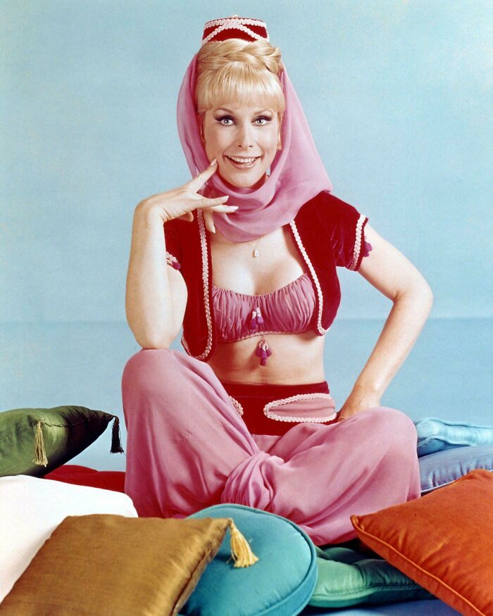 Actress in vintage pink and maroon costume sitting cross-legged on colorful pillows in a classic retro photo to feel old.