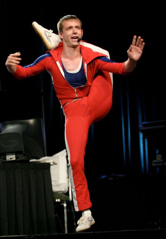 Flexible man performing high kick on stage wearing a red outfit, demonstrating proof that superhumans walk among us.