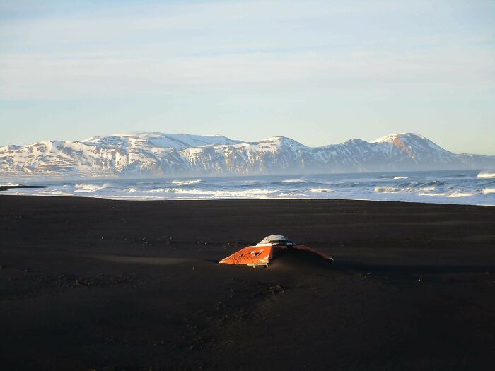 Partially buried orange object on a black sand beach with snowy mountains and ocean waves, interesting beach discovery.