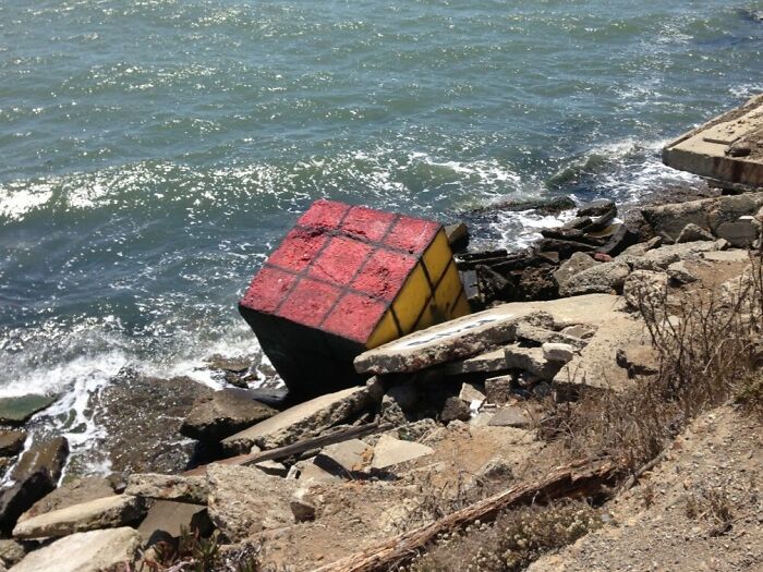 Large Rubik's Cube sculpture partially submerged among rocks by the beach, an interesting and bizarre find at the shore.