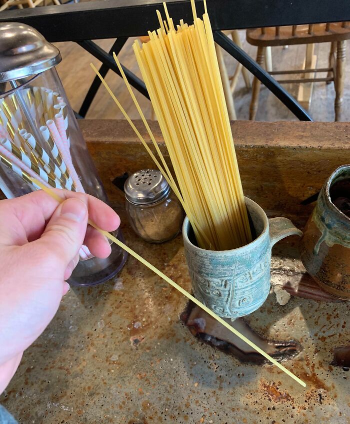 Hand holding uncooked spaghetti next to a container of pasta and a salt shaker in a creative bar or restaurant setting.