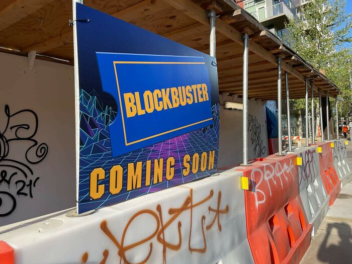 Construction site with a colorful Blockbuster sign and graffiti on barriers, highlighting random notes and messages outdoors.