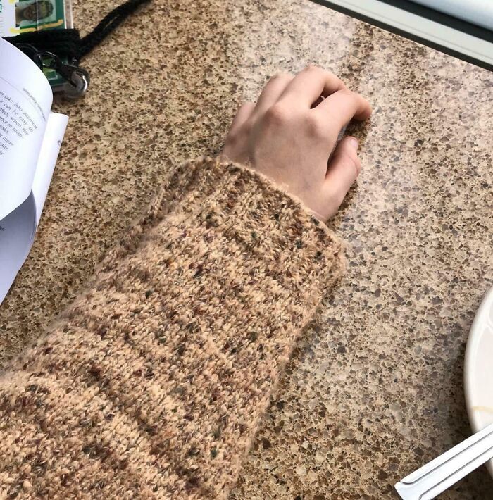 Hand wearing a brown knit sweater blending perfectly with the speckled brown tabletop, matching surroundings.