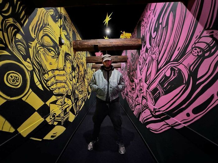 Colombian artist SakoAsko stands between two vibrant murals blending fantasy and reality in a narrow, dimly lit space.