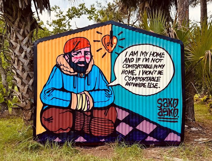 Mural by Colombian artist SakoAsko showing a seated man with a speech bubble about comfort and home.