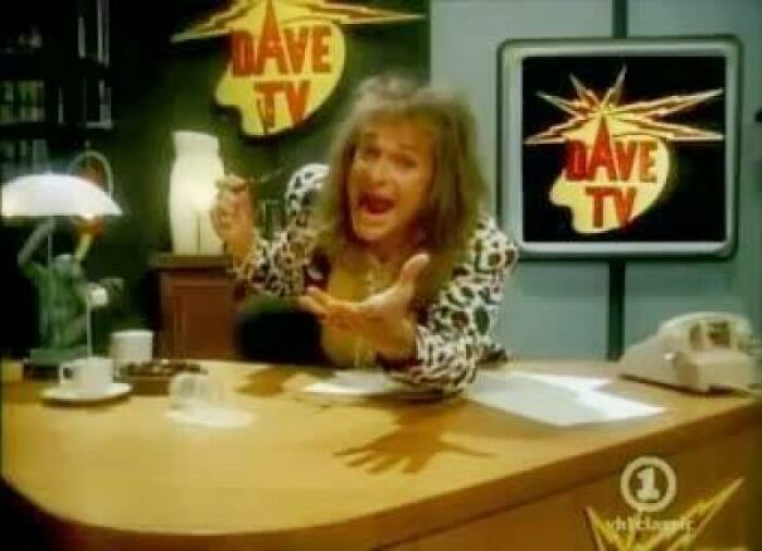 80s TV show host gesturing enthusiastically at a desk with vintage decor and a retro Dave TV logo in the background.