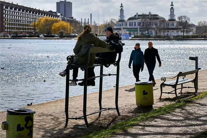 Two people sitting on an oversized Danish prison cell bench by the waterfront, with others walking nearby.