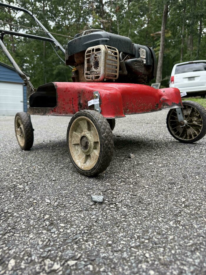 Homemade redneck engineer lawn mower with a modified rusty engine and worn wheels on an asphalt driveway outdoors.