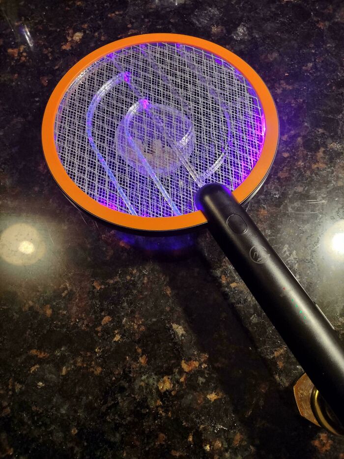 Electric bug zapper racket with glowing purple light resting on a dark granite surface, showcasing redneck engineer creativity.