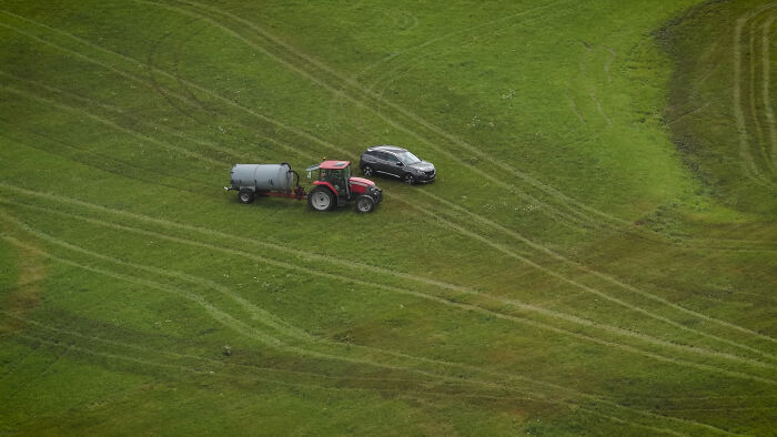 Aerial view of a farmer’s tractor spraying manure on a grassy field near a parked black car. Aerial view of a farmer’s tractor spraying manure on a grassy field near a parked black car.