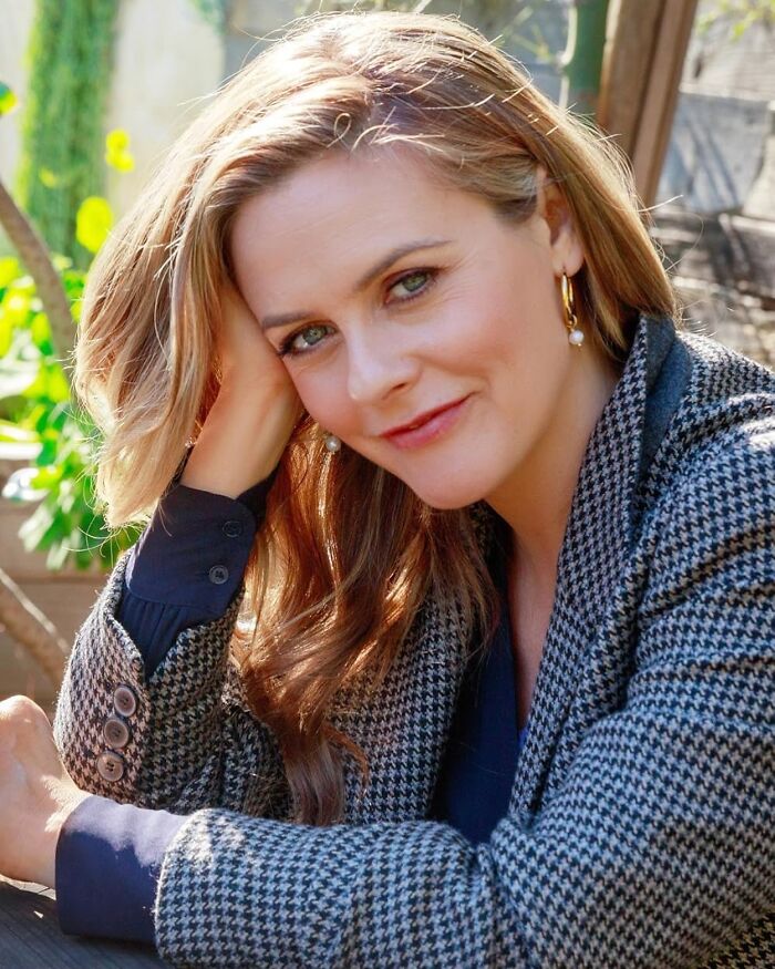 Alicia Silverstone smiling outdoors, wearing a patterned blazer and gold hoop earrings in natural daylight.