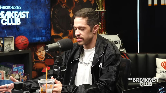Pete Davidson speaking into a microphone during a Hollywood celebrities interview on a radio show set.