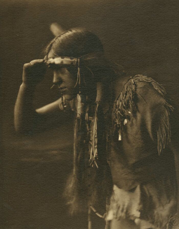 Sepia photograph of a Native tribesman from the 1900s wearing traditional attire and looking into the distance.