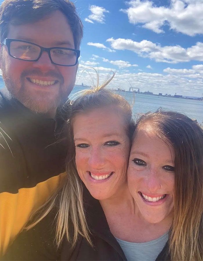 Conjoined twins Abby and Brittany Hensel smiling outdoors with a man by the water on a sunny day. Conjoined twins Abby and Brittany Hensel smiling outdoors with a man by the water on a sunny day.