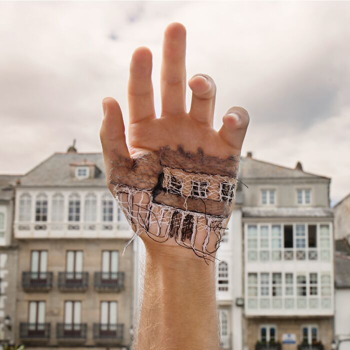 A hand with detailed stitched landscape artwork featuring a building, blending skin and fabric textures against a city backdrop.