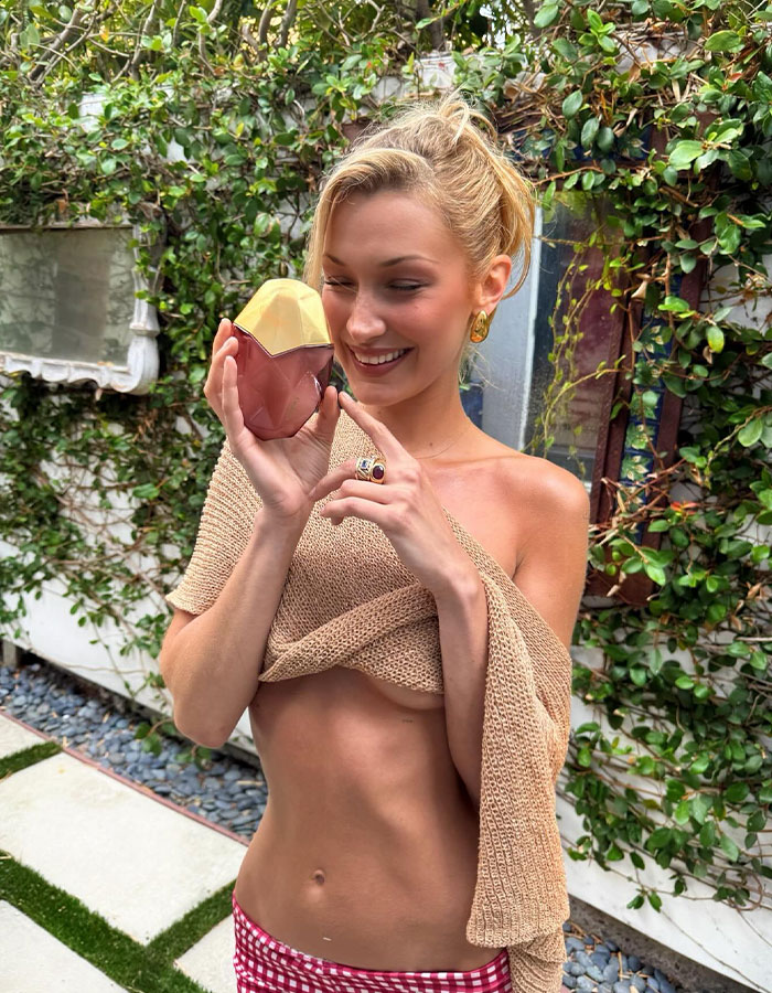 Bella Hadid showcasing a daring braless look while holding a perfume bottle in a lush outdoor setting. Bella Hadid showcasing a daring braless look while holding a perfume bottle in a lush outdoor setting.