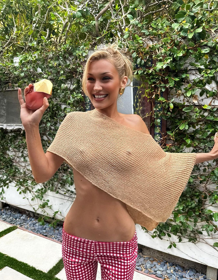 Bella Hadid in a daring braless look holding a perfume bottle outdoors with greenery in the background. Bella Hadid in a daring braless look holding a perfume bottle outdoors with greenery in the background.