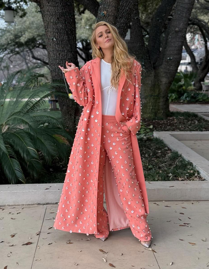 Blonde woman in a coral embellished coat and pants posing outdoors amid trees and greenery in a casual stance. Blonde woman in a coral embellished coat and pants posing outdoors amid trees and greenery in a casual stance.