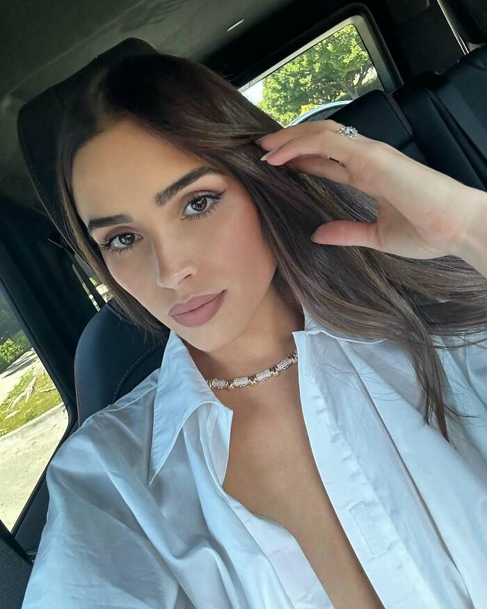 Miss Universe winner in a white shirt sitting inside a car, showcasing a diamond necklace and natural makeup look.