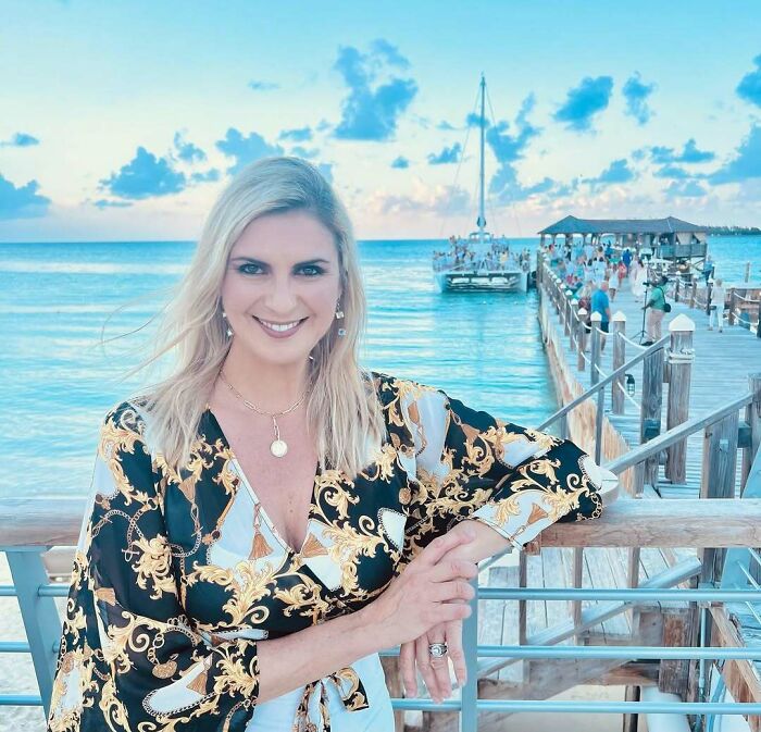 Blonde woman smiling by the ocean with a dock and boat in the background, showcasing Miss Universe winners style.
