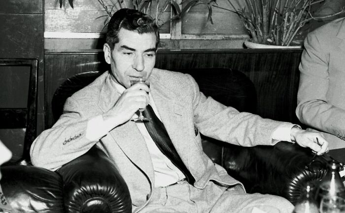 Black and white photo of a notorious mob boss in a suit sitting on a leather chair, drinking from a glass in a lounge setting.