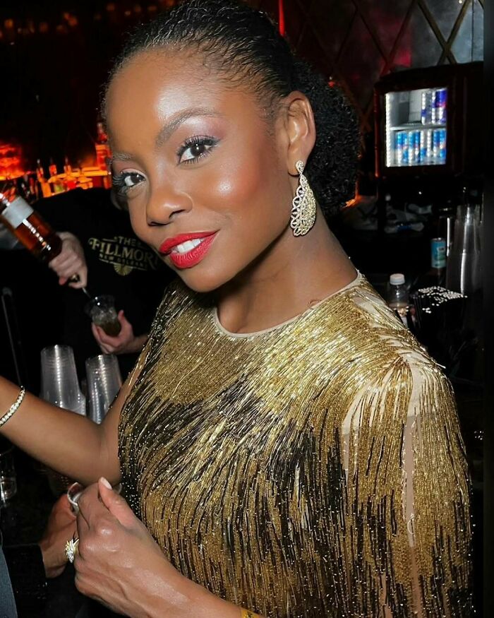 Miss Universe winner wearing gold sequined dress and chandelier earrings, smiling in a dimly lit elegant setting.