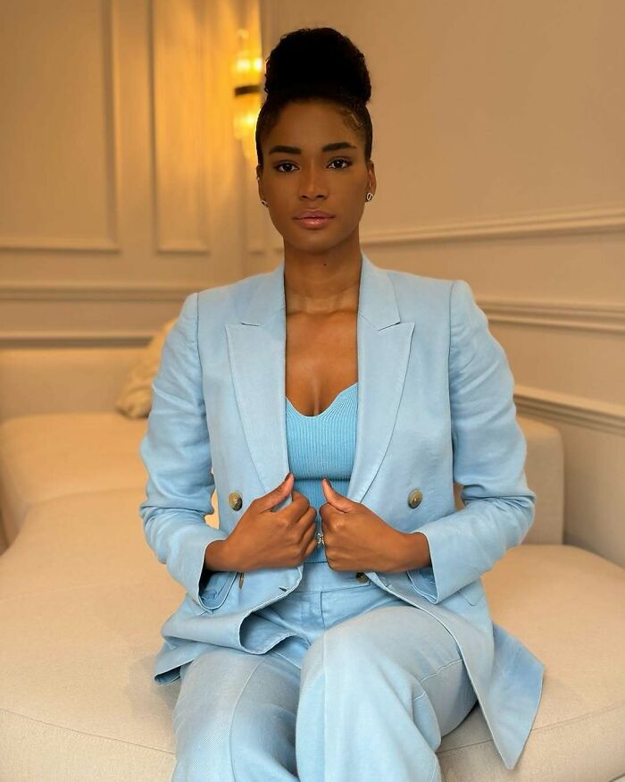 Miss Universe winner seated indoors wearing a light blue suit, showcasing elegant style and confident poise in a relaxed setting.