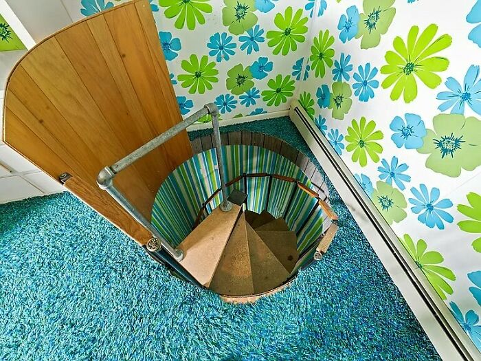 Spiral staircase with vintage door surrounded by colorful 1960s floral wallpaper and blue s**g carpet in a retro home.