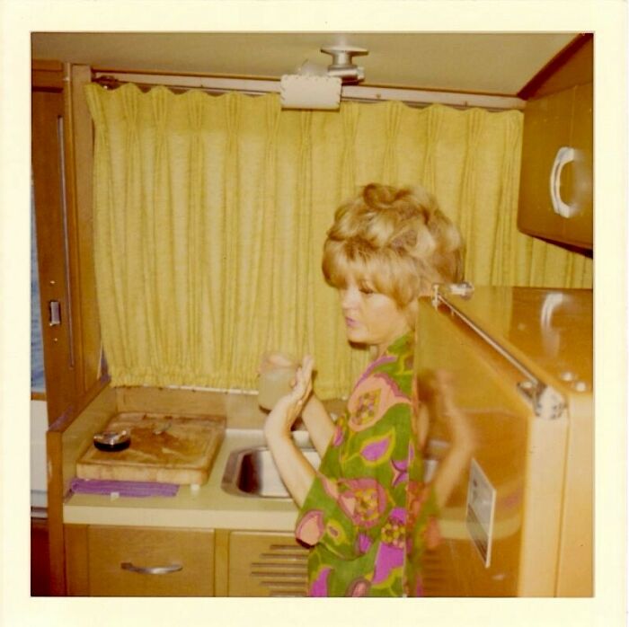 Woman in a vintage kitchen, holding a drink, surrounded by retro decor and yellow curtains, in June Cleavers vintage closet style.
