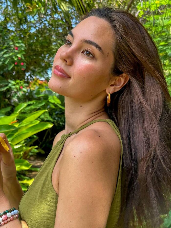 Miss Universe winner posing outdoors with natural makeup, wearing gold earrings and a green top in lush greenery.
