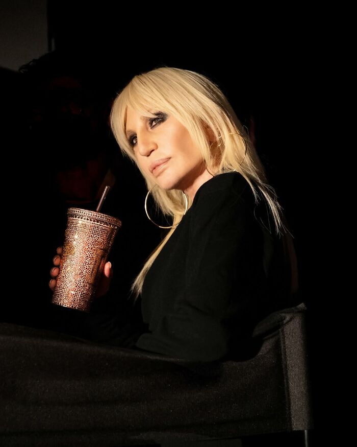 Donatella Versace with blonde hair and hoop earrings holding a sparkling cup, showcasing her stunning transformation. Donatella Versace with blonde hair and hoop earrings holding a sparkling cup, showcasing her stunning transformation.