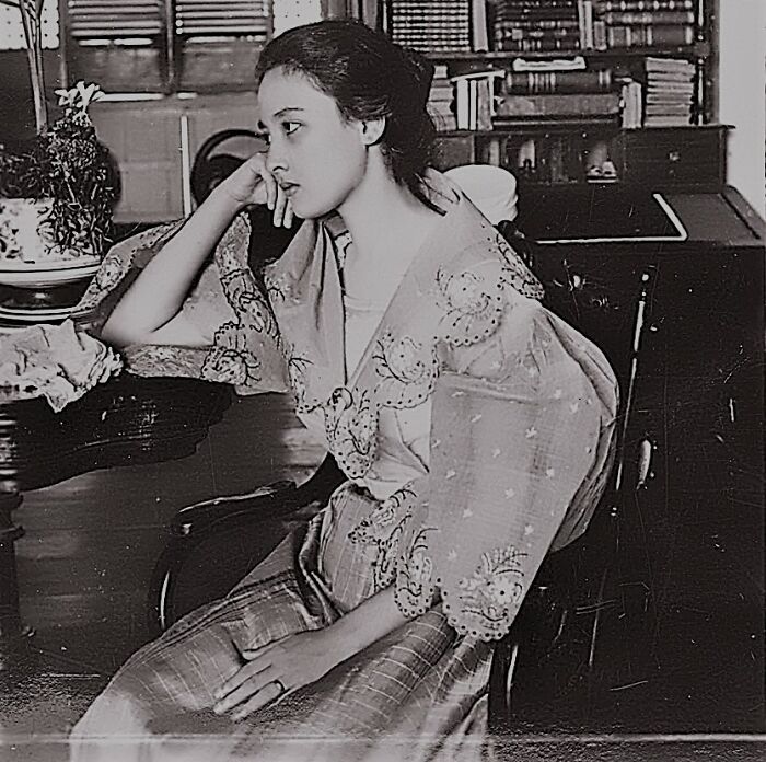Young Filipino woman in traditional 1890s clothing sitting thoughtfully by a table in a vintage room, Philippines 1890s.