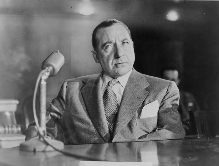 Black and white photo of a notorious mob boss in a suit sitting at a table with a vintage microphone in front of him.