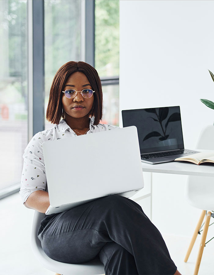 Young Gen Z worker wearing glasses and office attire, sitting with a laptop in a modern workplace setting. Young Gen Z worker wearing glasses and office attire, sitting with a laptop in a modern workplace setting.