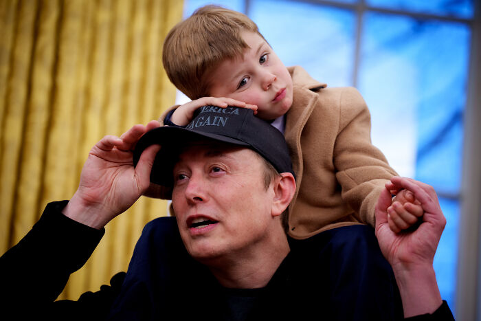 Elon Musk wearing a black cap with a young child on his shoulders in an indoor setting with blue and gold background. Elon Musk wearing a black cap with a young child on his shoulders in an indoor setting with blue and gold background.