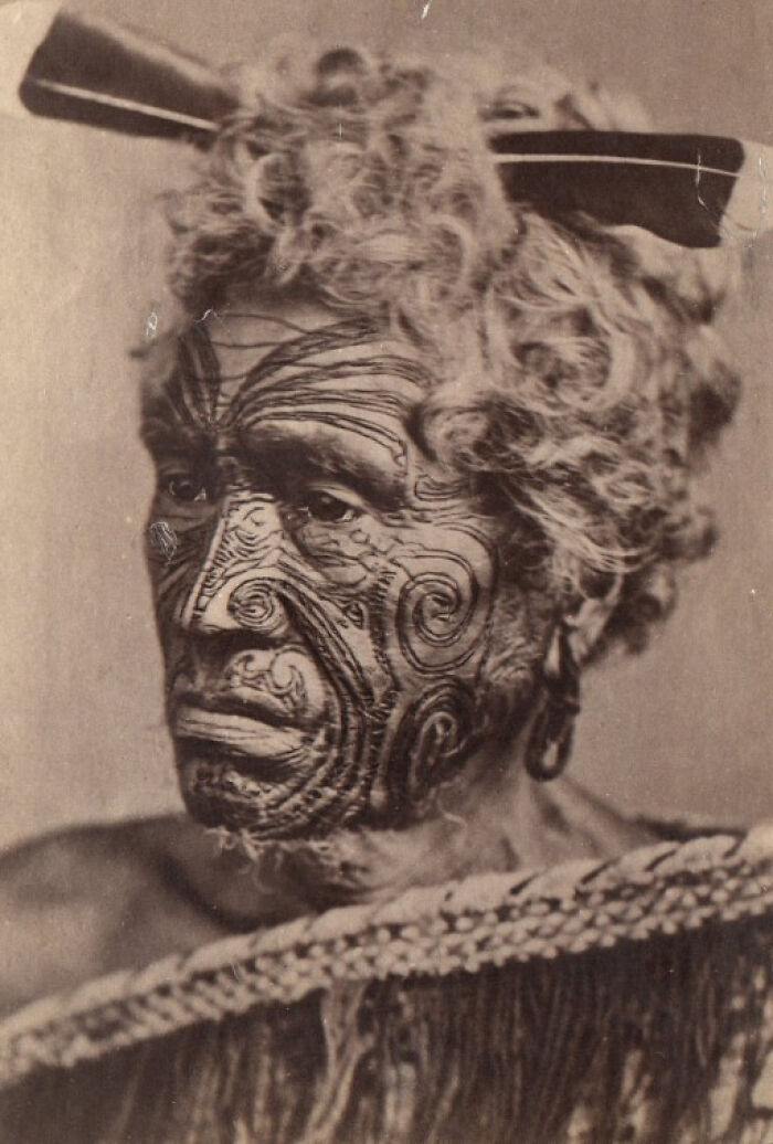 Elderly Māori man with traditional facial moko tattoo and curly hair wearing a woven cloak in a historic portrait.