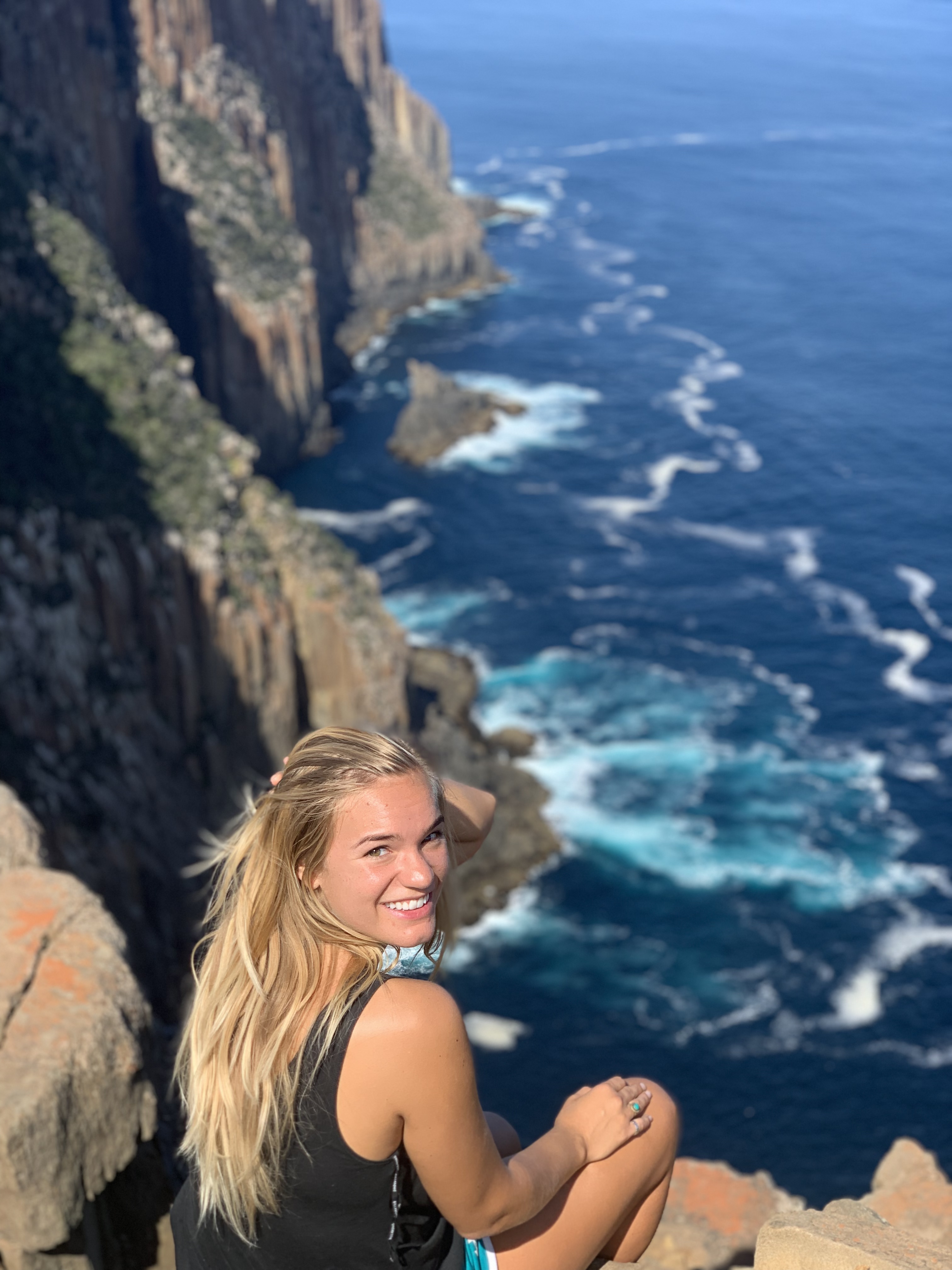 Young woman with blonde hair sitting on cliff edge overlooking ocean waves, representing one-way flight to Australia journey.