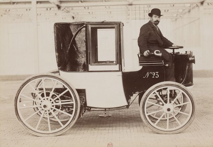 Early car with large spoked wheels and covered cabin, man with bowler hat seated, showing wild first cars from 100 years ago.