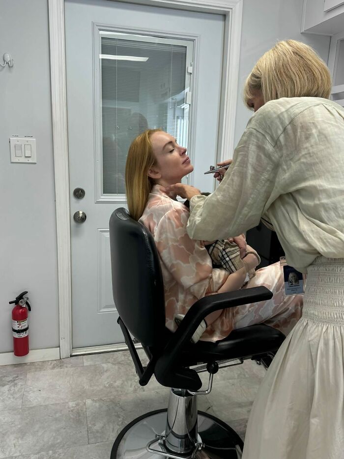 Lindsay Lohan seated in a chair, getting makeup applied, showcasing a natural, makeup-free look after plastic surgery rumors. Lindsay Lohan seated in a chair, getting makeup applied, showcasing a natural, makeup-free look after plastic surgery rumors.